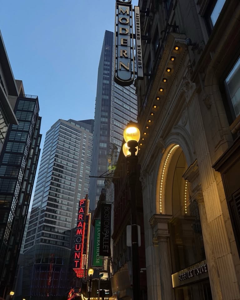 A city street view featuring the illuminated Modern Theatre and Suffolk University signs, with tall buildings and theater facades rising above the street.
