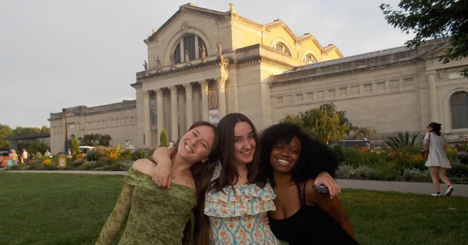 Three friends smile for a photo outdoors on a grassy lawn in front of a grand historic building, with landscaped gardens and other visitors in the background.