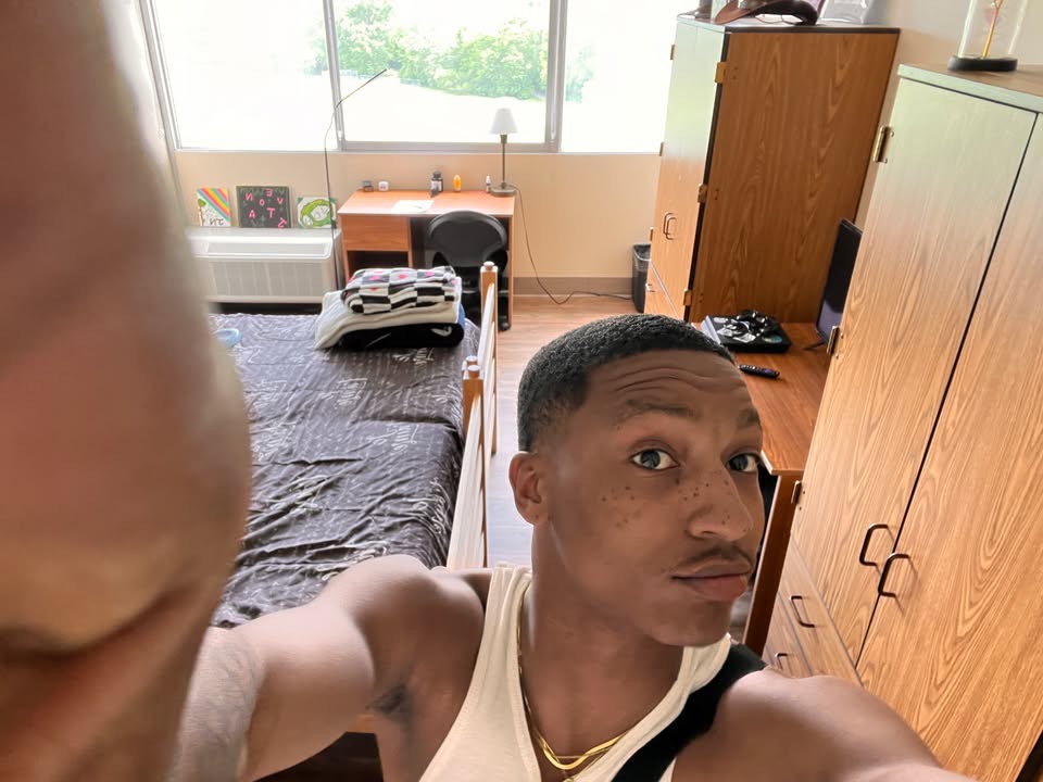 Javonte taking a selfie in his Towers room, with a neatly made bed, desk, chair, wardrobes, and large windows visible behind him
