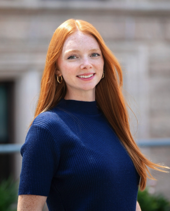 Lauren with long red hair smiles at the camera while wearing a navy blue top, with a blurred building exterior in the background