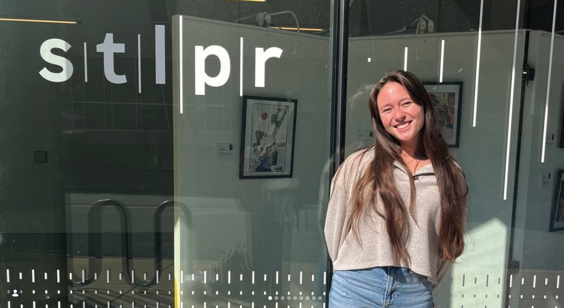 Madison with long brown hair smiles at the camera while standing in front of the studio for the St. Louis Public Radio Station