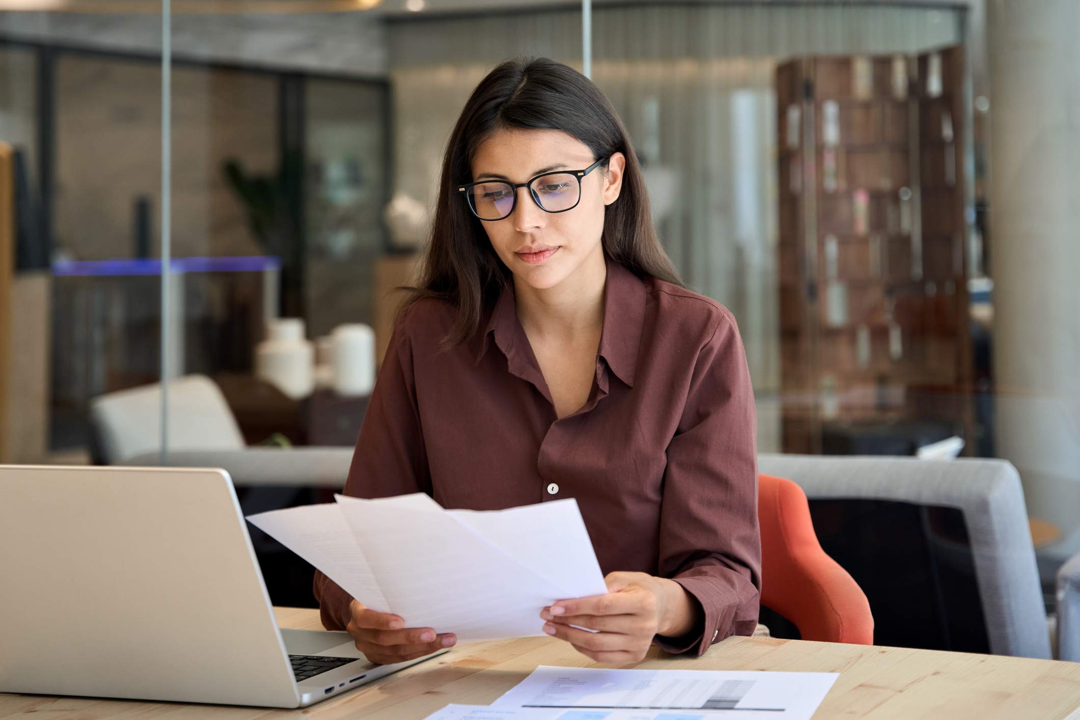 Business woman sitting at desk looking at papers with her laptop in front of her