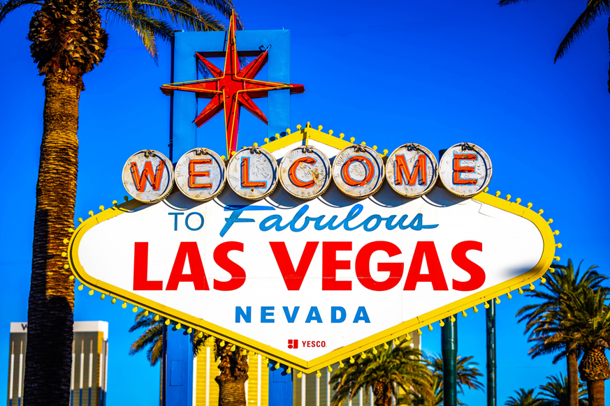  The iconic "Welcome to Fabulous Las Vegas Nevada" sign stands prominently under a clear, bright blue sky. The neon sign features a classic mid-century diamond shape with "Welcome" spelled out in individual circular letters across the top and "Las Vegas" in bold red lettering in the center. A large red starburst sits atop the blue support structure, and the scene is framed by tall palm trees on either side, with a glimpse of a resort building in the lower-left background.