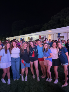 A large group of young adults pose together outdoors at night in front of a brightly lit building.