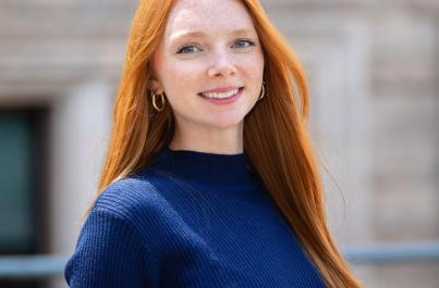 Lauren with long red hair smiles at the camera while wearing a navy blue top, with a blurred building exterior in the background