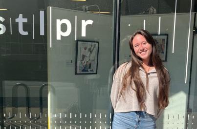 Madison with long brown hair smiles at the camera while standing in front of the studio for the St. Louis Public Radio Station