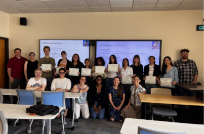 A group of students and faculty pose for a photo in a classroom, with several students holding certificates. Presentation screens displaying “2026 Research Teams” are visible in the background