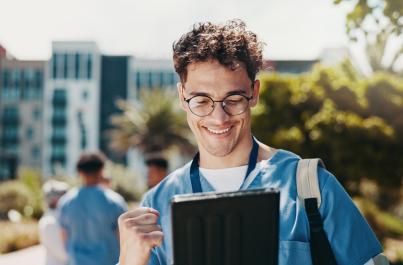 A young man wearing glasses and a blue shirt smiles while looking at a tablet on a university campus. He is wearing a backpack and lanyard, with campus buildings, trees, and other students in the background