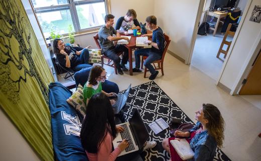 Young adults gather on couches and chairs. Three men are seated at a table. Everyone is smiling and studying.